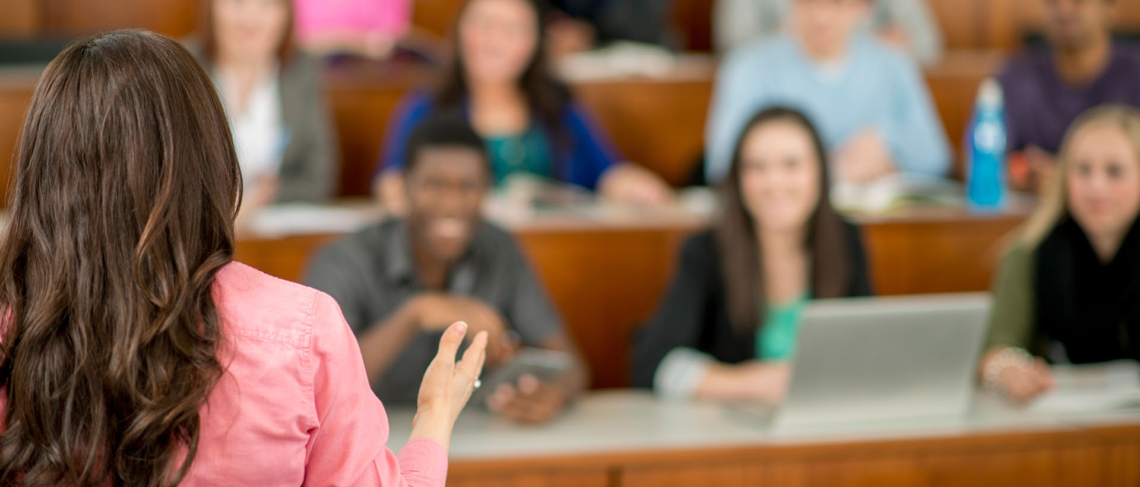 Imagen de una profesora de espaldas dando una clase a alumnos en un aula.
